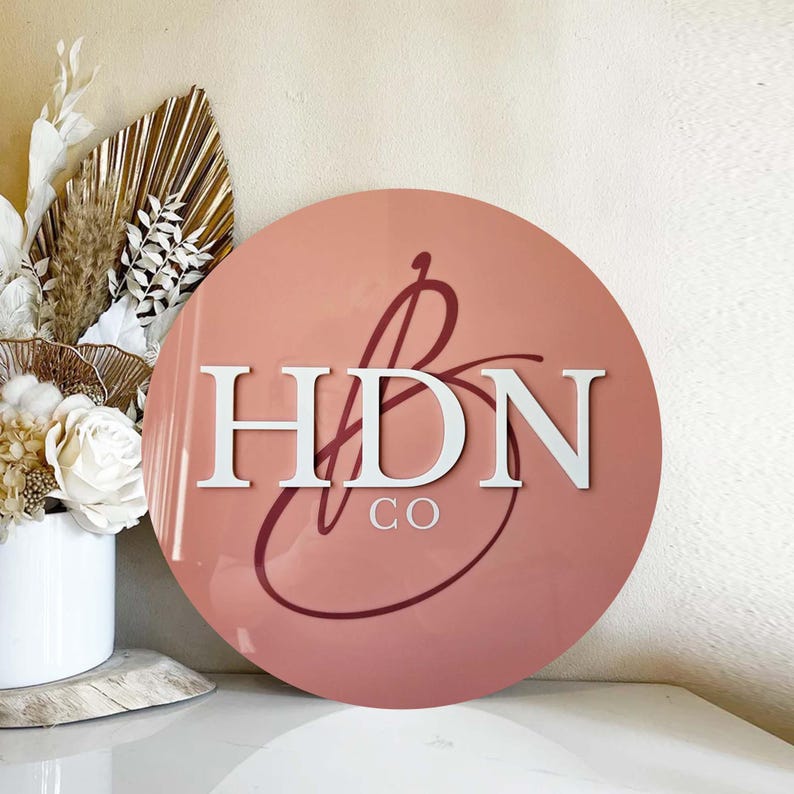 K&ouml;nnte beinhalten: Ein rundes, rosafarbenes Schild mit dem Text "HDN CO" in wei&szlig;en Buchstaben. Die Buchstaben sind mit einem kursiv geschriebenen "B" in dunklerem Rosa gestaltet.