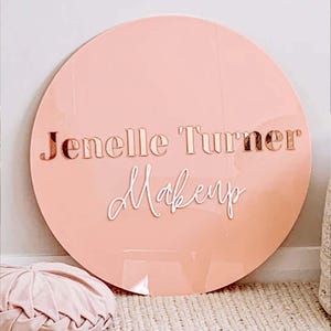 K&ouml;nnte beinhalten: Ein rundes, hellrosa Acrylschild mit dem Text "Jenelle Turner Makeup" in ros&eacute;goldenen und wei&szlig;en Buchstaben.