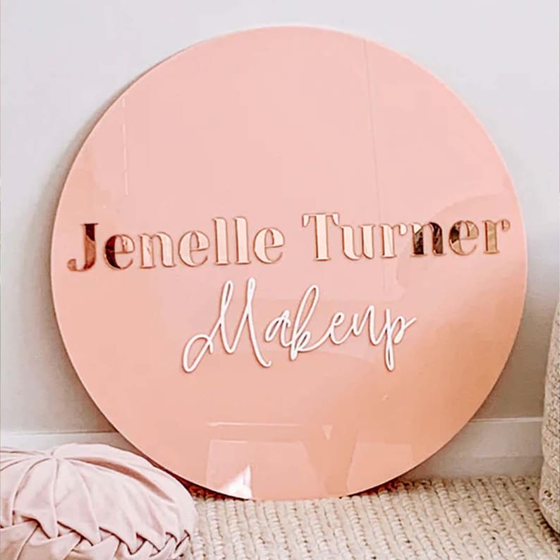 K&ouml;nnte beinhalten: Ein rundes, hellrosa Acrylschild mit dem Text "Jenelle Turner Makeup" in ros&eacute;goldenen und wei&szlig;en Buchstaben.