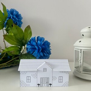 May include: A white paper model of a house with a brick facade and a tiled roof. The house has two windows on each side and a door in the centre. There are two small trees in front of the house. The model is sitting on a white surface.