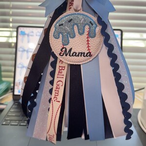 Peut inclure: Une d&eacute;coration de type mum sur le th&egrave;me du baseball, avec un n&oelig;ud bleu clair et des rubans en cascade. Le m&eacute;daillon central pr&eacute;sente un motif de baseball avec le mot "Mama". Des rubans bleu marine, blancs et bleu clair ajoutent &agrave; l'aspect festif.