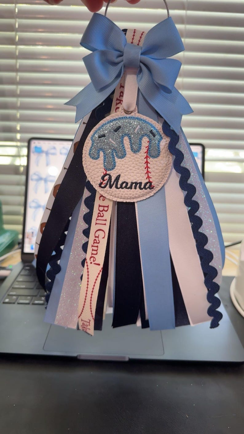 Peut inclure: Une d&eacute;coration de type mum sur le th&egrave;me du baseball, avec un n&oelig;ud bleu clair et des rubans en cascade. Le m&eacute;daillon central pr&eacute;sente un motif de baseball avec le mot "Mama". Des rubans bleu marine, blancs et bleu clair ajoutent &agrave; l'aspect festif.