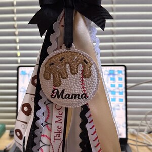 Peut inclure: Un porte-cl&eacute;s sur le th&egrave;me du baseball avec un n&oelig;ud noir et plusieurs rubans. Le charme central est un cercle blanc avec un motif de baseball et le mot "Mama". Les rubans incluent des coutures de baseball, des beignets et le texte "Take Me Out".
