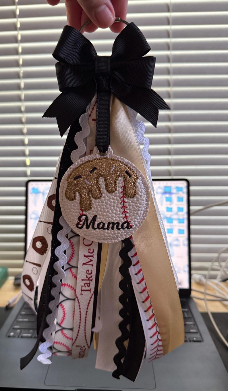 Peut inclure: Un porte-cl&eacute;s sur le th&egrave;me du baseball avec un n&oelig;ud noir et plusieurs rubans. Le charme central est un cercle blanc avec un motif de baseball et le mot "Mama". Les rubans incluent des coutures de baseball, des beignets et le texte "Take Me Out".