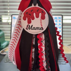 Peut inclure: D&eacute;coration sur le th&egrave;me du baseball avec un n&oelig;ud rouge en haut. La d&eacute;coration pr&eacute;sente un cercle blanc avec des accents rouges et le mot "Mama". Des rubans rouges, noirs et blancs descendent.
