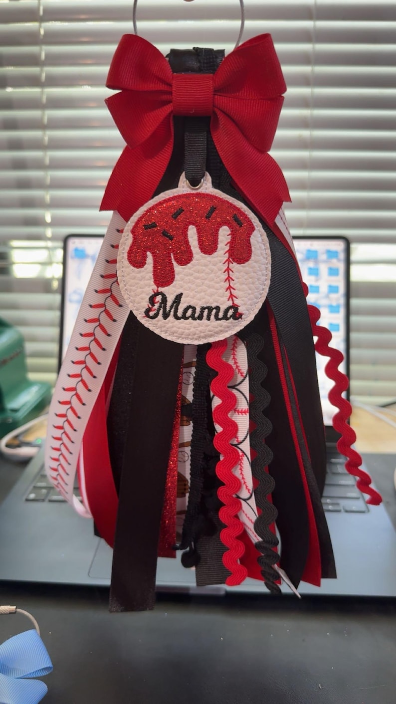 Peut inclure: D&eacute;coration sur le th&egrave;me du baseball avec un n&oelig;ud rouge en haut. La d&eacute;coration pr&eacute;sente un cercle blanc avec des accents rouges et le mot "Mama". Des rubans rouges, noirs et blancs descendent.
