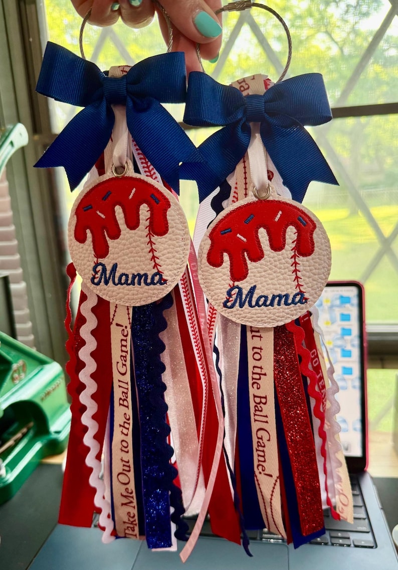 Peut inclure: Deux porte-cl&eacute;s sur le th&egrave;me du baseball avec des rubans et des n&oelig;uds rouges, blancs et bleus. Chaque porte-cl&eacute;s pr&eacute;sente un motif de baseball avec du gla&ccedil;age rouge et le mot "Mama". Les rubans incluent le texte "Take Me Out to the Ball Game!"