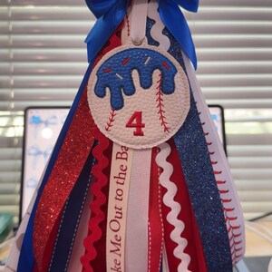 Peut inclure: D&eacute;coration sur le th&egrave;me du baseball avec un n&oelig;ud bleu et des rubans rouges, blancs et bleus en cascade. La pi&egrave;ce ma&icirc;tresse pr&eacute;sente un motif de baseball avec le chiffre 4. Le texte "Take Me Out to the Ba" est visible sur un ruban.