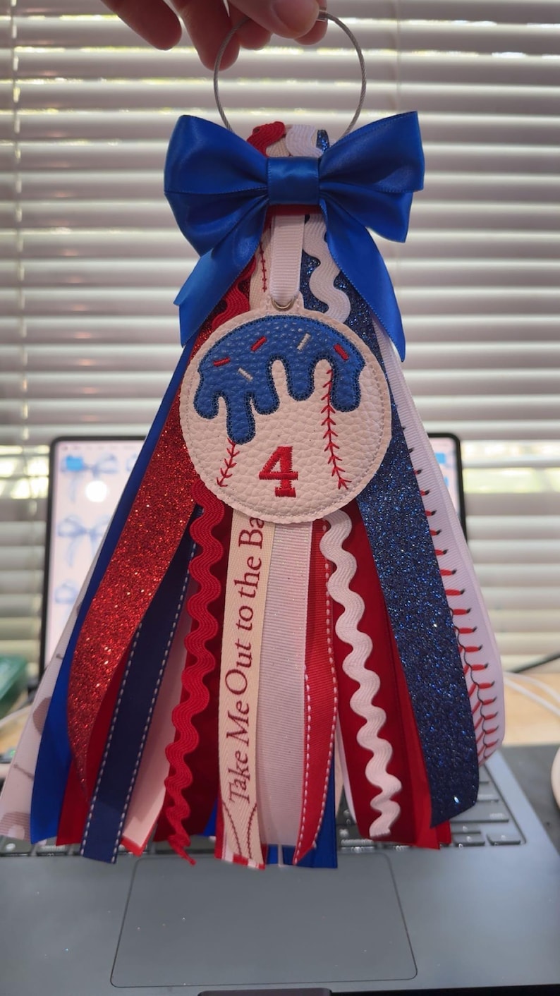 Peut inclure: D&eacute;coration sur le th&egrave;me du baseball avec un n&oelig;ud bleu et des rubans rouges, blancs et bleus en cascade. La pi&egrave;ce ma&icirc;tresse pr&eacute;sente un motif de baseball avec le chiffre 4. Le texte "Take Me Out to the Ba" est visible sur un ruban.
