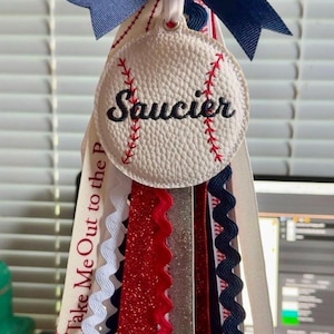 May include: A baseball-themed mum with a white, textured baseball-shaped centerpiece embroidered with the name "Saucier" in black. Red stitching details the baseball. Navy blue bow and ribbons in red, white, and navy blue complete the design.