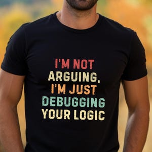 May include: Black crew neck t-shirt with the text "I'M NOT ARGUING, I'M JUST DEBUGGING YOUR LOGIC" in a retro font. The text is in red, orange, teal, and yellow. The shirt is worn by a person.