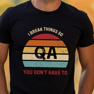 May include: Black t-shirt with a retro sunset design. The design includes the text "I BREAK THINGS SO QA YOU DON'T HAVE TO" in white and the letters "QA" in a large font. The shirt is short-sleeved.