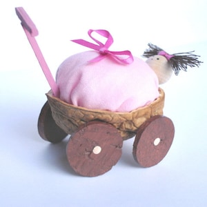 May include: A pink fabric doll in a walnut shell baby carriage with pink ribbon and a pink handle.