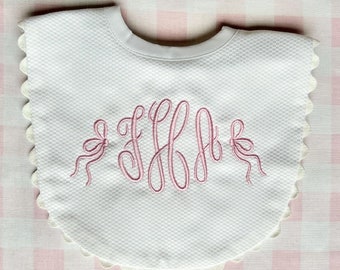 May include: White baby bib with a textured waffle weave, scalloped edges, and a pink embroidered monogram. Two pink bows flank the monogram. The bib has a white satin neckline.