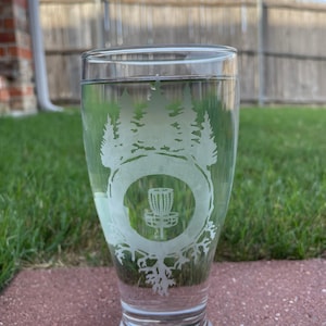 May include: A clear glass beer mug with an etched design of a disc golf basket surrounded by trees. The design is white and the mug is filled with a clear liquid.