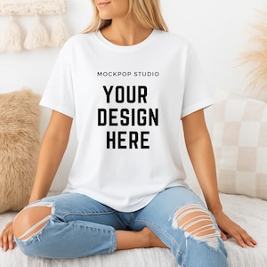 Op de afbeelding: Een wit T-shirt met de tekst "MOCKPOP STUDIO YOUR DESIGN HERE" gedrukt op de voorkant. Het T-shirt wordt gedragen door een persoon die op een witte bank zit.