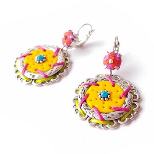 Puede incluir: Un par de pendientes hechos a mano con un diseño de filigrana plateada. Los pendientes presentan un centro amarillo con un acento turquesa, costuras rosas y verdes y una parte superior rosa y naranja. Tienen cierre de palanca.
