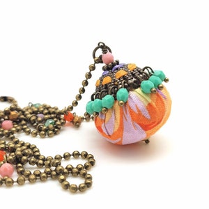May include: A colorful fabric pendant necklace with a bronze chain and turquoise beads. The pendant is a round ball with a swirl pattern in orange, pink, and purple.