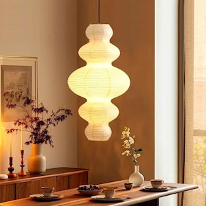 May include: A white paper pendant light with a multi-tiered, rounded, and stacked silhouette. The lamp features a ribbed texture and emits a warm, soft glow, suspended from a thin black cord above a wooden dining table set with various items.