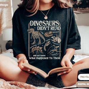 May include: Dark gray t-shirt with a dinosaur-themed graphic. The shirt features illustrations of dinosaur skeletons and skulls, along with the text "DINOSAURS DIDN'T READ" and "and we know What Happened To Them". A black mug with "PNG FILE ONLY" and "DIGITAL DOWNLOAD" text is also visible.