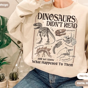 May include: Beige sweatshirt with a dinosaur-themed graphic. The graphic includes dinosaur skeletons and the text "DINOSAURS DIDN'T READ and we know What Happened To Them". The design features a T-Rex, Velociraptor, Triceratops, and other dinosaurs.