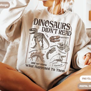 May include: Cream-colored sweatshirt featuring a dinosaur-themed graphic. The design includes dinosaur skeletons and illustrations with the text "DINOSAURS DIDN'T READ" and "and we know What Happened To Them". A digital download is available.