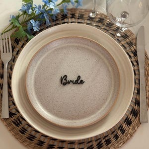 Op de afbeelding: Een gedekte tafel voor een bruiloft met een beige gespikkelde bord, een kleiner bord en een geweven placemat. Het woord "Bride" van zwart hout ligt op het bord. Een vork, een mes en een wijnglas zijn ook aanwezig.