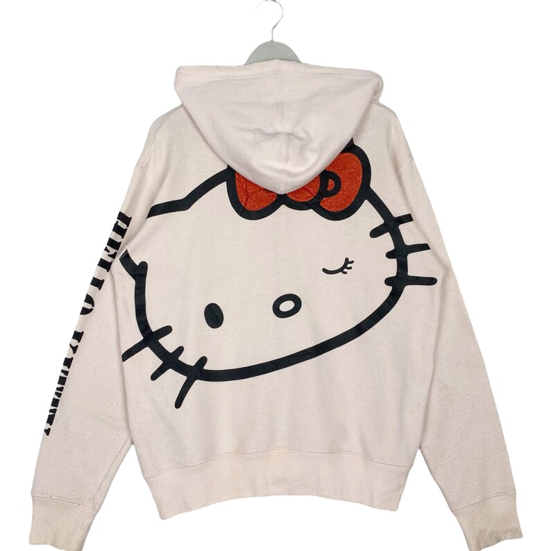 May include: A cream-colored hooded sweatshirt featuring a large Hello Kitty graphic on the back. The graphic includes a red bow and black outlines. The left sleeve has black text. The sweatshirt is hanging on a hanger.