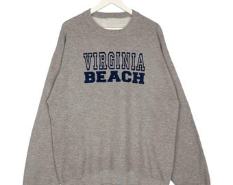 Puede incluir: Sudadera gris con la inscripción "VIRGINIA BEACH" en letras azul marino en el pecho. La sudadera tiene cuello redondo y mangas largas, y está colgada de una percha blanca.