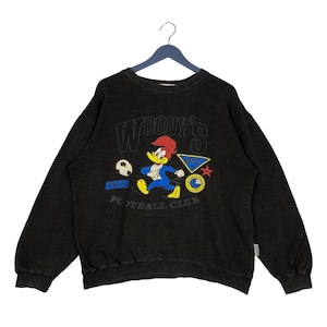 Op de afbeelding: Zwart sweatshirt met een cartoon van Woody Woodpecker die voetbal speelt. Het sweatshirt heeft de tekst "Woody's Football Club" erop gedrukt.