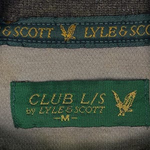 Puede incluir: Primer plano de una etiqueta de camisa gris y verde. La etiqueta dice "CLUB L/S by LYLE & SCOTT -M-" con un emblema de &aacute;guila dorada. La parte superior de la camisa tiene una etiqueta verde con las palabras "LYLE & SCOT" y un emblema de &aacute;guila dorada.