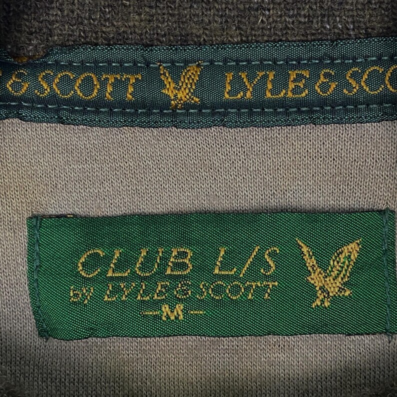 Puede incluir: Primer plano de una etiqueta de camisa gris y verde. La etiqueta dice "CLUB L/S by LYLE & SCOTT -M-" con un emblema de &aacute;guila dorada. La parte superior de la camisa tiene una etiqueta verde con las palabras "LYLE & SCOT" y un emblema de &aacute;guila dorada.