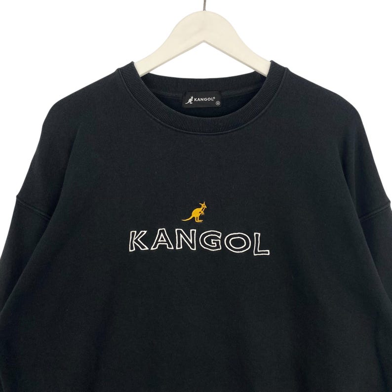 Puede incluir: Sudadera negra con el nombre de la marca "KANGOL" en letras blancas delineadas. Un logotipo de canguro amarillo est&aacute; encima del nombre de la marca. La sudadera est&aacute; colgada de una percha blanca.