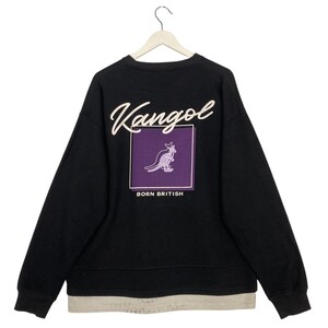 May include: Black sweatshirt with the Kangol logo in white script across the top. A purple square with a kangaroo graphic and the words "BORN BRITISH" are centered on the back. The sweatshirt has a layered hem.