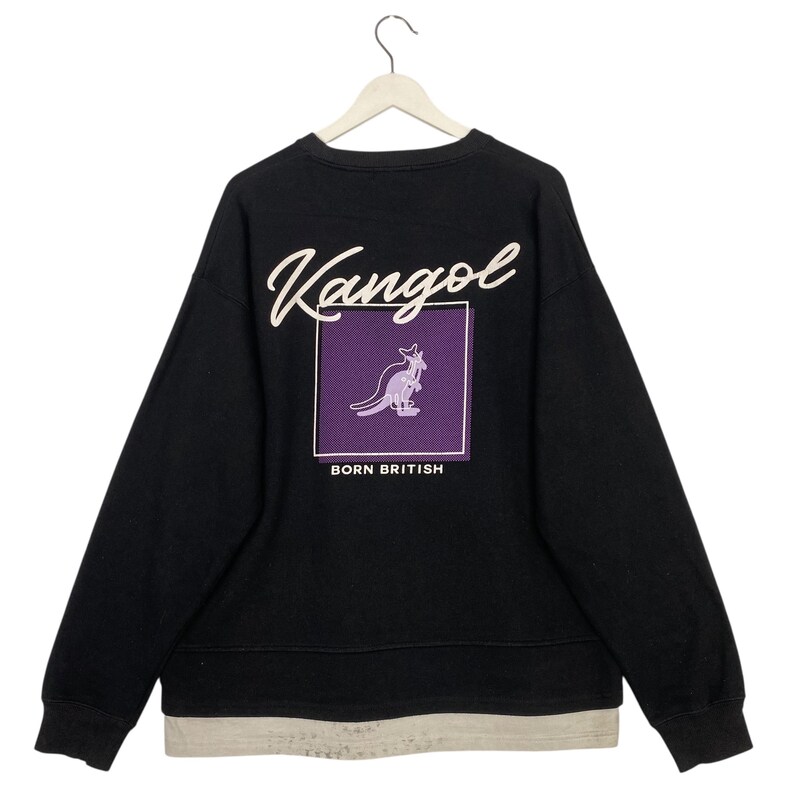 May include: Black sweatshirt with the Kangol logo in white script across the top. A purple square with a kangaroo graphic and the words "BORN BRITISH" are centered on the back. The sweatshirt has a layered hem.