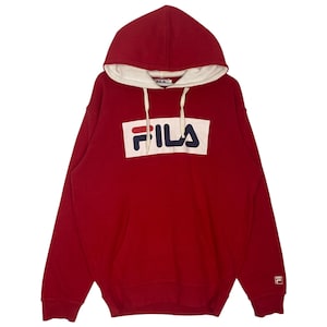 May include: A red hooded sweatshirt with the FILA logo in a white rectangle on the front. The hood has a cream-colored lining. The sweatshirt has a kangaroo pocket and a small FILA logo on the left sleeve.