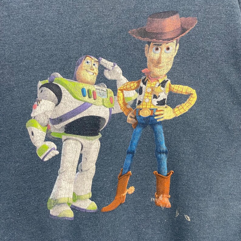 Op de afbeelding: Marineblauw grafisch T-shirt met Buzz Lightyear en Woody uit Toy Story. Buzz Lightyear draagt een wit en groen ruimtepak en Woody draagt een cowboyhoed, vest en een blauwe jeans.