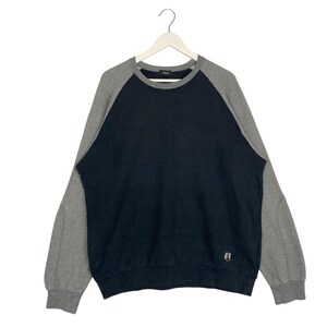 Puede incluir: Una sudadera de color bloque negro y gris con un pequeño logotipo en la parte inferior izquierda. La sudadera tiene cuello redondo y mangas largas.