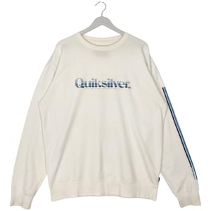 May include: Off-white sweatshirt with the Quiksilver logo in blue and white across the chest. A blue and white stripe runs down the left sleeve. The sweatshirt is hanging on a gray hanger.