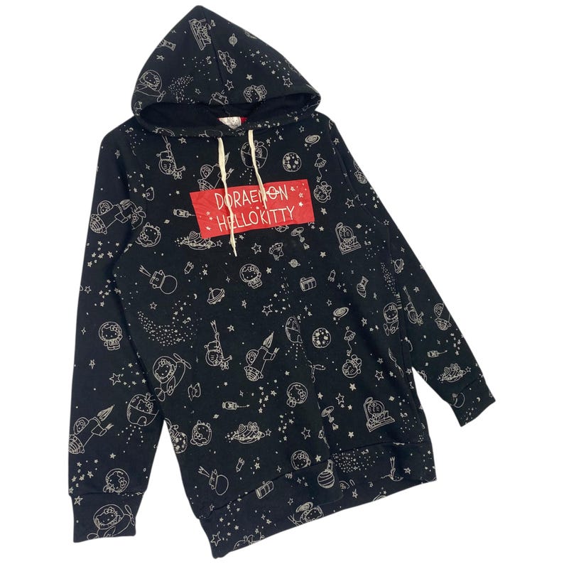 K&ouml;nnte beinhalten: Schwarzer Kapuzenpullover mit einem sich wiederholenden Muster aus Zeichentrickfiguren, Planeten und Sternen. Der Pullover hat einen roten rechteckigen Aufn&auml;her mit den wei&szlig;en Worten "Doraemon Hello Kitty". Die Kapuze hat einen Kordelzug.