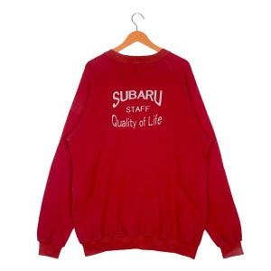 May include: A red crew neck sweatshirt with the text "SUBARU STAFF Quality of Life" in white. The sweatshirt is hanging on a wooden hanger.