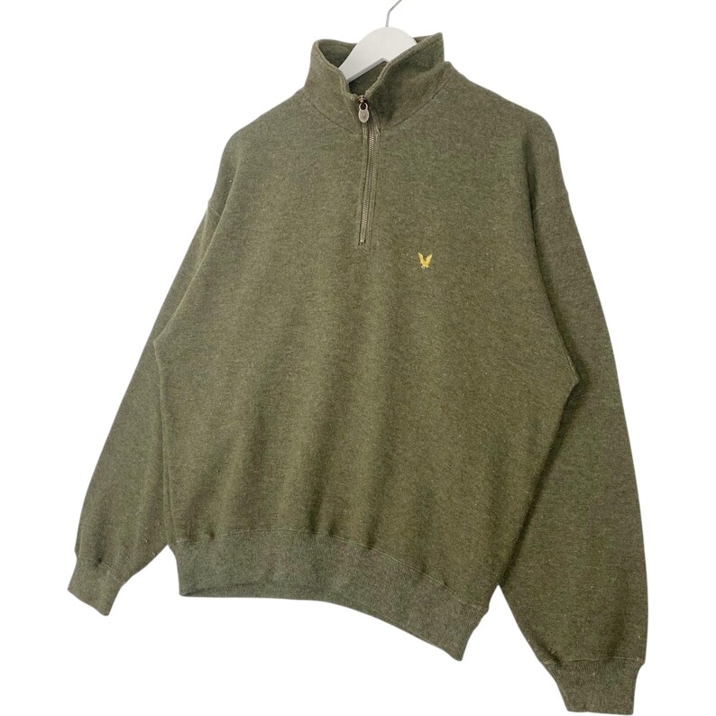 Puede incluir: Sudadera verde oliva con cierre de cremallera de un cuarto y un peque&ntilde;o logotipo amarillo en el pecho. La parte superior de manga larga est&aacute; hecha de un material suave y tiene dobladillo y pu&ntilde;os acanalados. La sudadera est&aacute; colgada en una percha blanca.