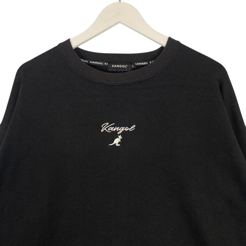May include: Black crew neck sweatshirt with the brand name "Kangol" embroidered in white script on the chest. The brand name is also printed around the neckline. The sweatshirt is hanging on a white hanger.