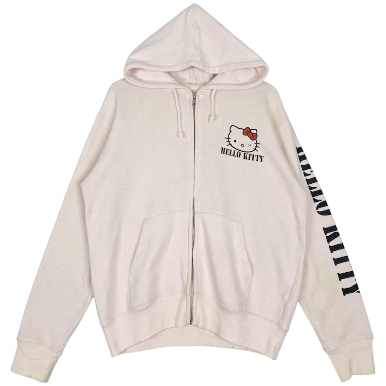 May include: Cream-colored zip-up hoodie featuring a Hello Kitty graphic with a red bow and the text "HELLO KITTY". The right sleeve displays the text "HELLO KITTY" in black. The hoodie has a hood and front pockets.