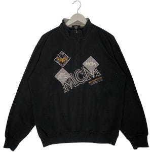 Puede incluir: Sudadera negra MCM con cuello con cremallera. La sudadera presenta el logotipo de MCM en forma de diamante, junto con otros diseños en forma de diamante. También es visible el texto "MODE CREATION MUNCHEN".