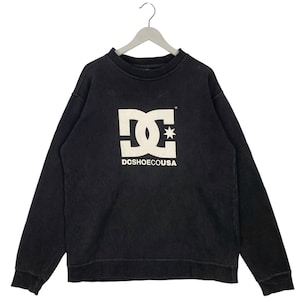 May include: Black crew neck sweatshirt with the white DC Shoes logo and text "DCSHOECOUSA". The logo features a stylized "DC" design with a star. The sweatshirt is hanging on a white hanger.