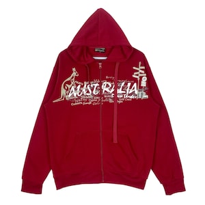 Puede incluir: Sudadera con capucha roja con cremallera, un canguro y la palabra "AUSTRALIA" impresa en la parte delantera. La sudadera tiene una capucha con cordón y un bolsillo canguro.