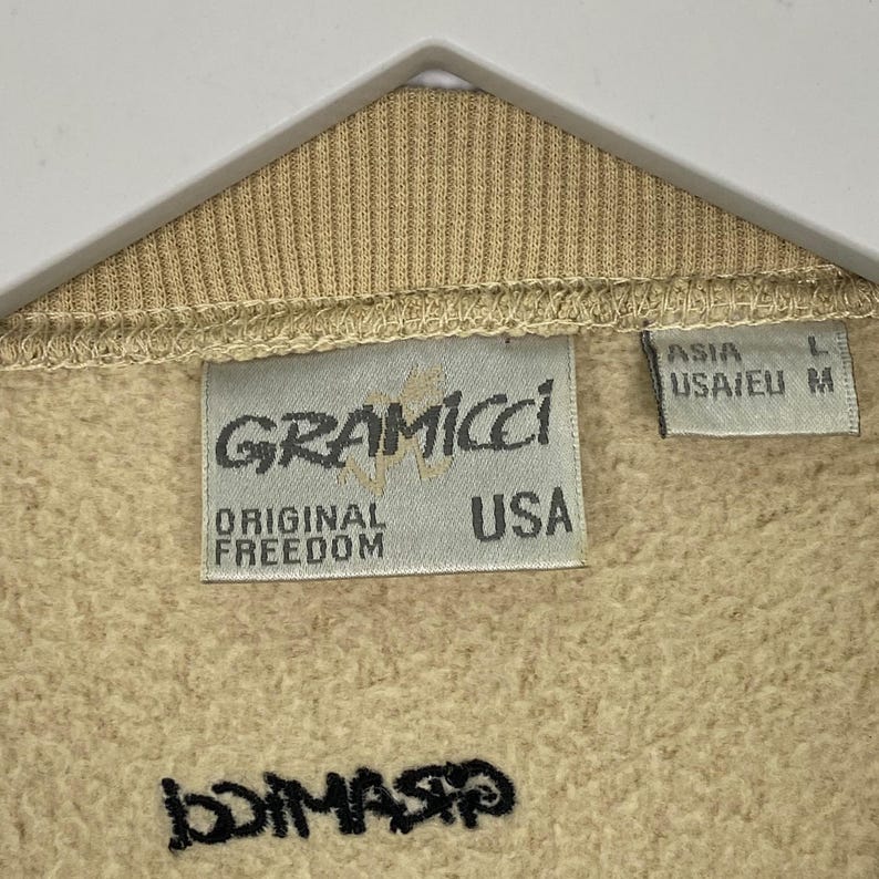 May include: Close-up of a tan Gramicci sweatshirt. The label reads "GRAMICCI ORIGINAL FREEDOM USA". The size tag reads "ASIA L USA/EU M". The bottom of the sweatshirt has the word "DIMASIO" printed in black.