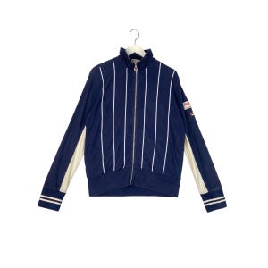 Puede incluir: Una chaqueta azul marino con cremallera con rayas verticales blancas y ribetes blancos en las mangas. La chaqueta tiene un logotipo "FILA" en la manga derecha.