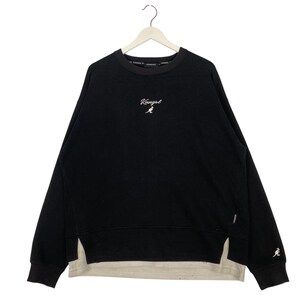 May include: Black sweatshirt with the white "Kangol" logo and kangaroo emblem. The sweatshirt has a layered design with a light beige underlayer. The long-sleeved top is hanging on a white hanger.